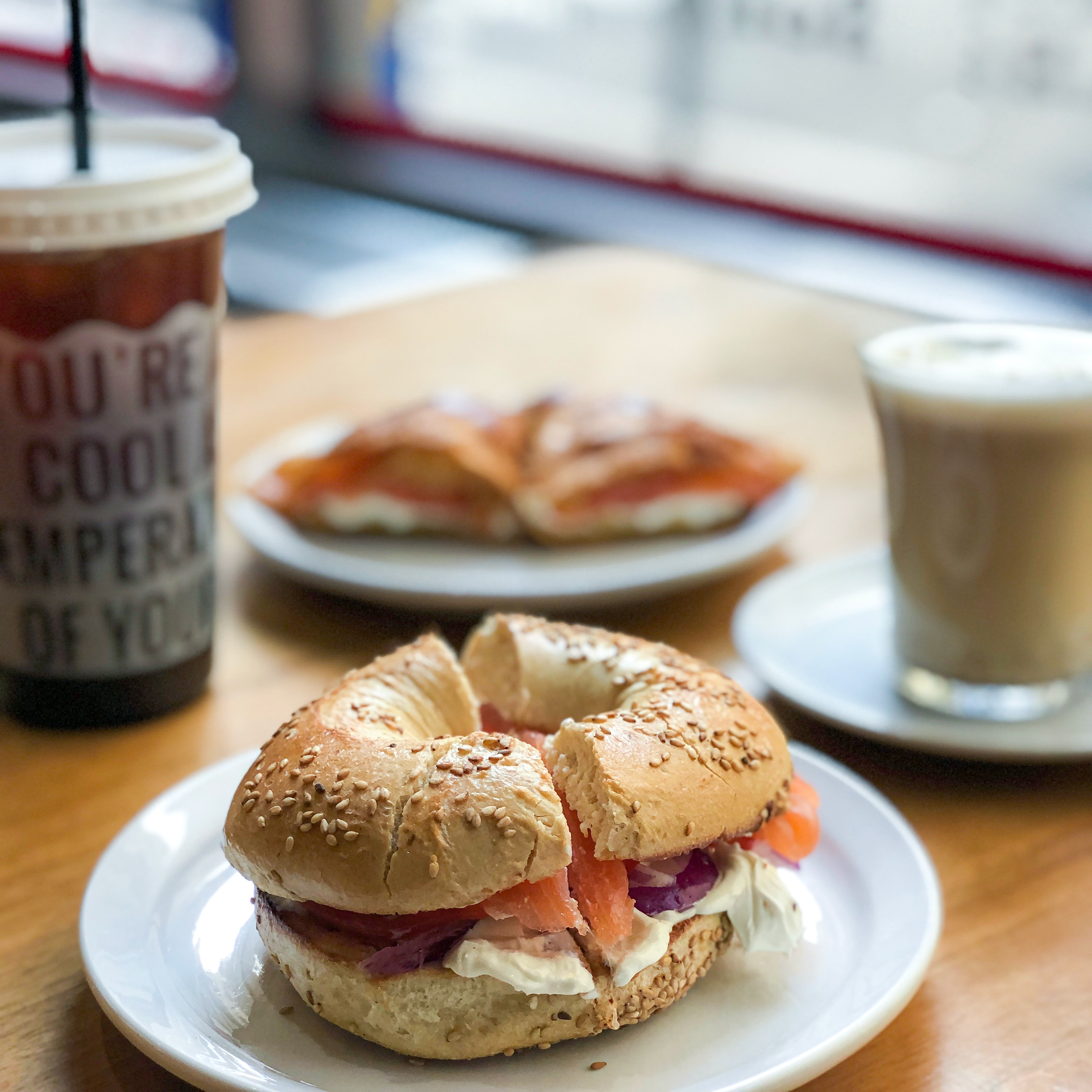 plated smoked salmon, cream cheese and red onion on a sesame seed bagel on a wooden table. Iced coffee and latte blurred in the background